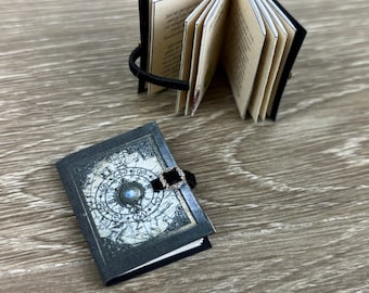 May include: Miniature open book with tan pages and black cover, next to a closed miniature book. The closed book has a dark blue cover with a celestial design and a black ribbon closure. The open book is held together with a black elastic band.