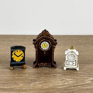 May include: Three miniature decorative clocks. The clocks are in black, brown, and white. The black clock has a yellow clock face. The brown clock has a gold-colored clock face. The white clock has a silver-colored clock face.