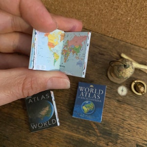 Puede incluir: Dos atlas mundiales en miniatura, uno abierto en un mapa del mundo y el otro cerrado con el título "World Atlas & Encyclopedia" visible. También se ve un pequeño globo terráqueo y una brújula en la imagen.