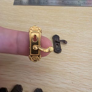 Könnte beinhalten: Ein goldfarbener Miniatur-Türgriff mit aufwendigen Details wird gezeigt. Ein Lineal zeigt Messungen in Zoll. Ein kleinerer, dunkel gefärbter Türgriff befindet sich neben dem goldenen. Mehrere andere Miniatur-Türgriffe sind unten im Bild.