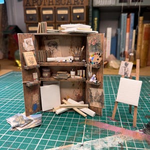 May include: A miniature wooden art studio cabinet with open doors, featuring shelves stocked with tiny books, paint tubes, brushes, and canvases. A small wooden easel stands to the right, holding a blank white canvas. The interior is decorated with art prints.