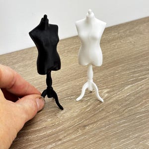 Puede incluir: Maniquíes en miniatura en blanco y negro. La forma negra está sostenida por una mano. Las formas están sobre un soporte de tres patas. La forma blanca está a la derecha. La forma negra está a la izquierda.