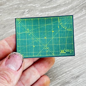 May include: A small, rectangular green cutting mat with yellow grid lines and a dark green border. The mat features various angle markings and measurements. The item is held in a person's hand.