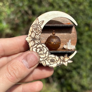 May include: A wooden crescent moon shelf with floral carvings, featuring two shelves. The top shelf holds a small, ornate, amber-coloured jar with a lid. The bottom shelf holds two clear quartz crystals.