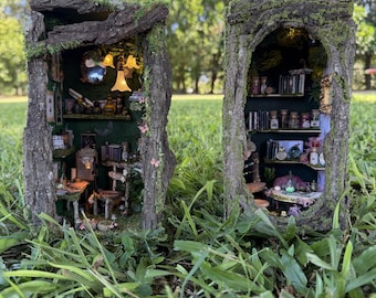 May include: Two miniature dioramas housed within hollowed-out tree bark structures covered in green moss. Each interior features multiple levels with tiny furniture, books, glass bottles, and small hanging light fixtures that emit a warm glow.