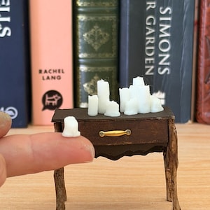 May include: A small, brown wooden table with a drawer and a gold-coloured handle, holding several white candles. The table has twig-like legs. A small white candle sits on a finger. Books are in the background.
