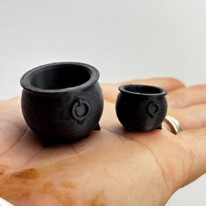 May include: Two small, black cauldrons with rounded bodies, three small feet, and a ring on the side. The cauldrons are displayed on a hand, with a light background. The cauldrons are likely used for decorative purposes or as miniature props.