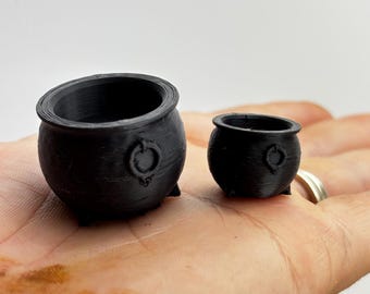 Op de afbeelding: Twee kleine, zwarte ketels met afgeronde lichamen, drie kleine voetjes en een ring aan de zijkant. De ketels worden op een hand gepresenteerd, met een lichte achtergrond. De ketels worden waarschijnlijk gebruikt voor decoratieve doeleinden of als miniatuur rekwisieten.