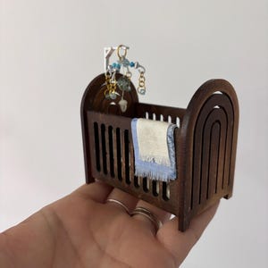 Puede incluir: Una cuna de madera en miniatura con una manta blanca y azul y un móvil con cuentas azules y doradas colgando encima.