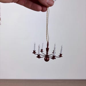 Può includere: Lampadario in miniatura con otto luci bianche a forma di candela. Il lampadario è rosso scuro con una catena e cablaggio dorati. Il lampadario è tenuto da una mano su sfondo bianco.