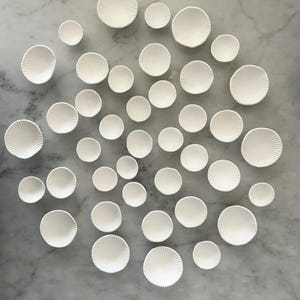 Puede incluir: Vista a&eacute;rea de una colecci&oacute;n de peque&ntilde;os cuencos de cer&aacute;mica blanca dispuestos sobre una superficie de m&aacute;rmol. Los cuencos var&iacute;an en tama&ntilde;o y presentan un patr&oacute;n radial estriado. La disposici&oacute;n es dispersa, creando una exhibici&oacute;n visualmente interesante.