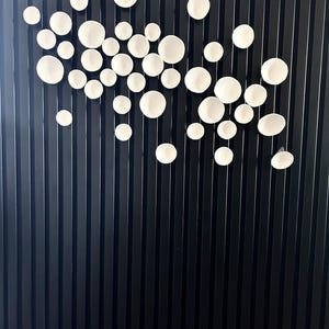 Puede incluir: Una instalaci&oacute;n de arte mural con numerosos elementos circulares blancos de cer&aacute;mica texturizada de varios tama&ntilde;os. Las piezas est&aacute;n dispuestas sobre un fondo oscuro con l&iacute;neas verticales, creando una exhibici&oacute;n moderna y art&iacute;stica. Los c&iacute;rculos blancos parecen flotar.