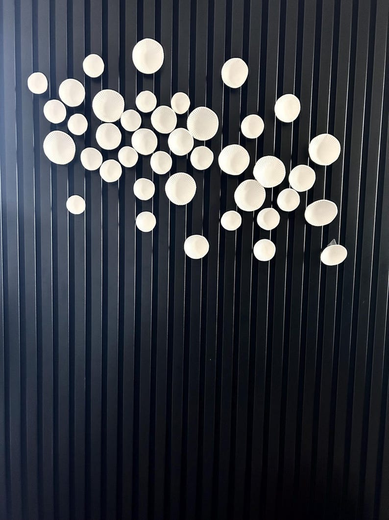 Puede incluir: Una instalaci&oacute;n de arte mural con numerosos elementos circulares blancos de cer&aacute;mica texturizada de varios tama&ntilde;os. Las piezas est&aacute;n dispuestas sobre un fondo oscuro con l&iacute;neas verticales, creando una exhibici&oacute;n moderna y art&iacute;stica. Los c&iacute;rculos blancos parecen flotar.