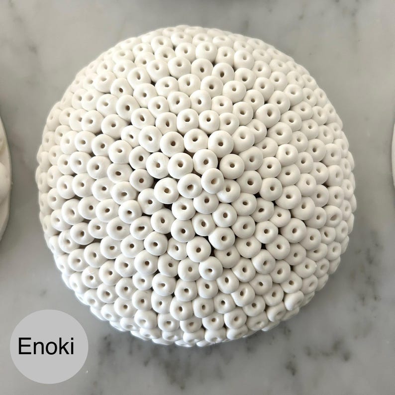May include: A white, spherical decorative object covered in small, donut-shaped elements. The surface is textured with numerous repeating forms. The word "Enoki" is printed in a white rectangle in the lower left corner.
