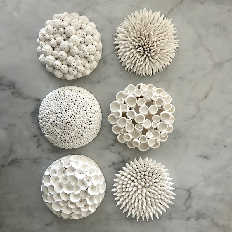 May include: Six white ceramic decorative wall art pieces with various textures. The pieces are round, resembling coral or sea urchins, with surfaces of small spheres, spikes, and open cylinders. The art is displayed on a marble surface.