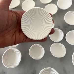 Puede incluir: Una mano sostiene un plato redondo de cer&aacute;mica blanca con un dise&ntilde;o radial texturizado. Varios platos similares est&aacute;n dispersos sobre una superficie clara, creando un patr&oacute;n. Los platos tienen una forma poco profunda y c&oacute;ncava.