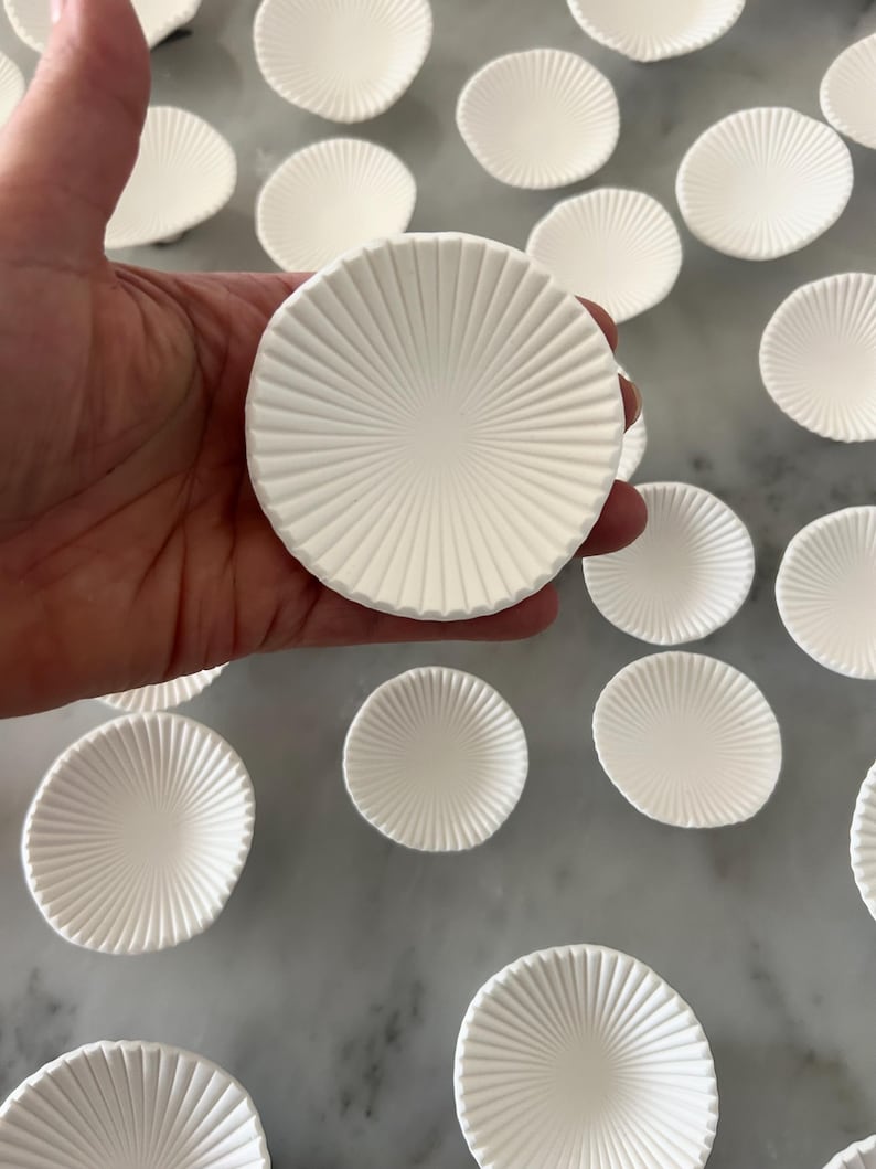 Puede incluir: Una mano sostiene un plato redondo de cer&aacute;mica blanca con un dise&ntilde;o radial texturizado. Varios platos similares est&aacute;n dispersos sobre una superficie clara, creando un patr&oacute;n. Los platos tienen una forma poco profunda y c&oacute;ncava.