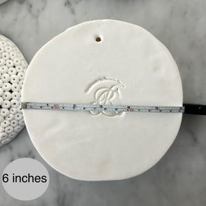 May include: A round, white ceramic piece with a hole at the top. A measuring tape shows the diameter is 6 inches. The piece is stamped with the words "Handmade by Rosaria Conte" and a stylized "R" design.