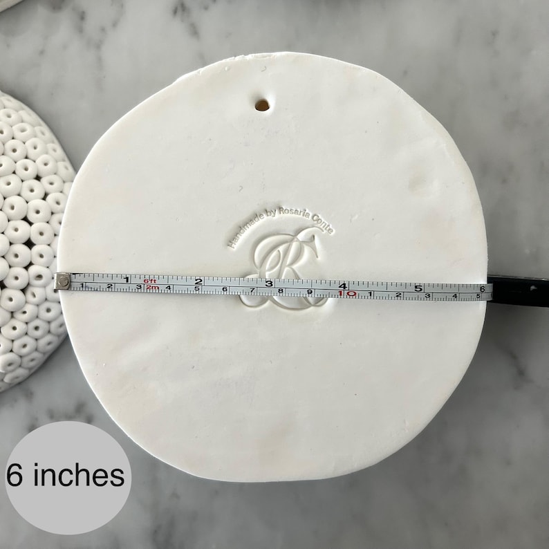 May include: A round, white ceramic piece with a hole at the top. A measuring tape shows the diameter is 6 inches. The piece is stamped with the words "Handmade by Rosaria Conte" and a stylized "R" design.