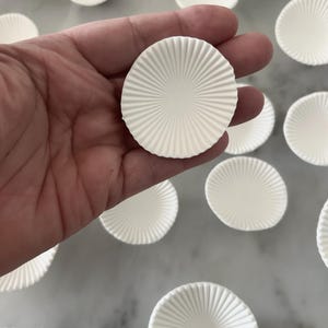 Puede incluir: Primer plano de varios moldes redondos de papel blanco para hornear con un dise&ntilde;o acanalado. Una taza se sostiene en una mano, mientras que otras est&aacute;n dispersas sobre una superficie de color claro. Las tazas tienen un patr&oacute;n radial que emana del centro.