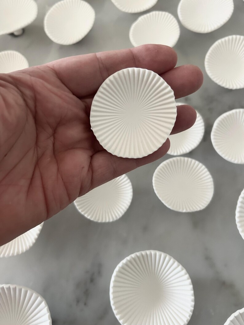 Puede incluir: Primer plano de varios moldes redondos de papel blanco para hornear con un dise&ntilde;o acanalado. Una taza se sostiene en una mano, mientras que otras est&aacute;n dispersas sobre una superficie de color claro. Las tazas tienen un patr&oacute;n radial que emana del centro.