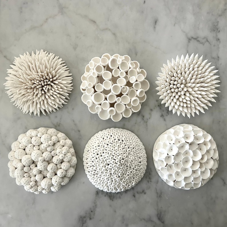 May include: Six white ceramic decorative pieces with various textures, including spiky, bubbly, and porous designs. The pieces are arranged on a light gray surface, showcasing their intricate details and organic forms. These pieces are ideal for home decor.
