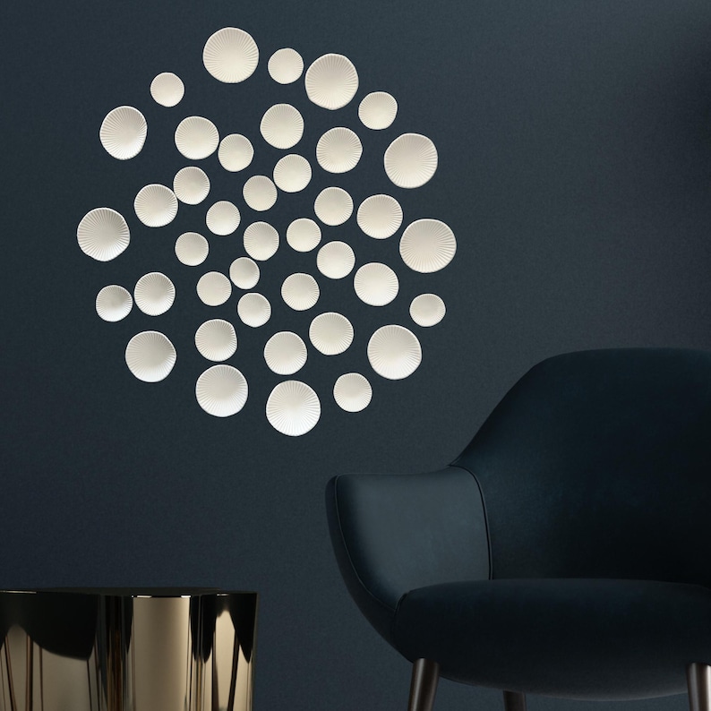 Puede incluir: Un conjunto de decoraciones murales circulares blancas y texturizadas de diferentes tama&ntilde;os dispuestas en un patr&oacute;n circular contra una pared azul oscuro. Un sill&oacute;n azul oscuro y una mesa auxiliar dorada est&aacute;n en primer plano.
