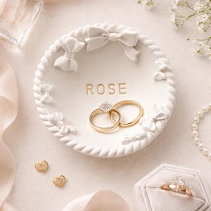 May include: A white ceramic ring dish with the word "ROSE" in gold lettering, adorned with bow and floral details. Two gold wedding rings with a diamond are inside the dish. Other items include a perfume bottle, a ring box, and pearl jewelry.