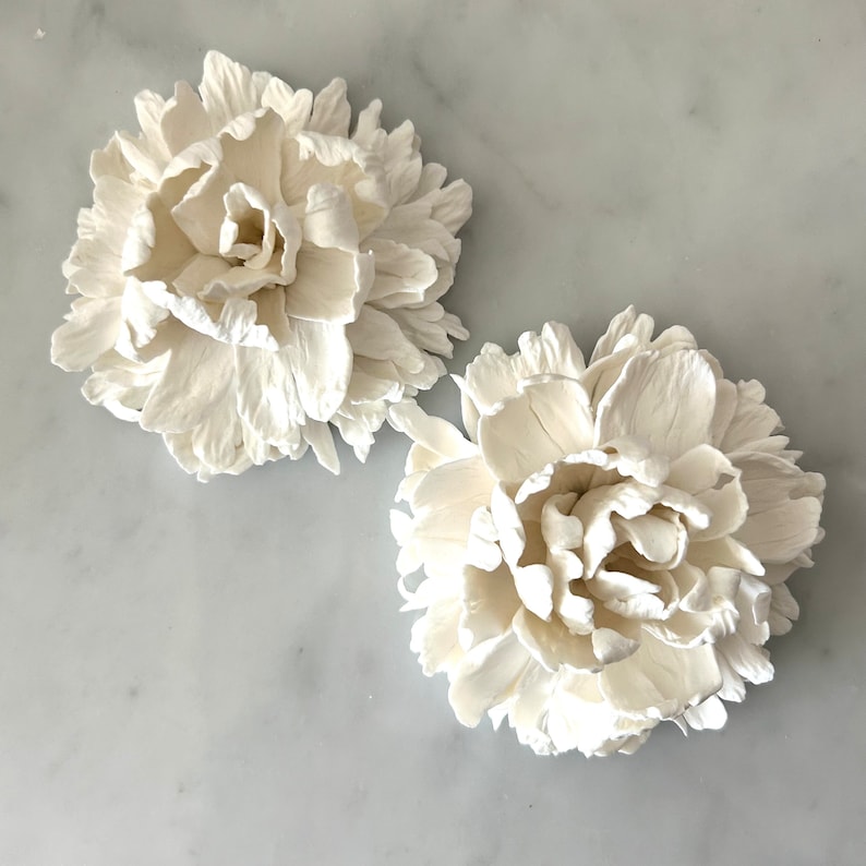 May include: Two white ceramic peonies, each with multiple layers of petals, are arranged on a white marble surface.