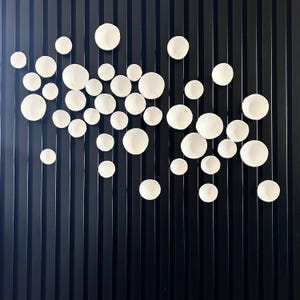 Puede incluir: Una instalaci&oacute;n de arte mural que presenta numerosos elementos decorativos circulares blancos y texturizados de varios tama&ntilde;os. Las piezas est&aacute;n dispuestas sobre un fondo oscuro con l&iacute;neas verticales, creando una est&eacute;tica moderna y minimalista. Las piezas blancas tienen un patr&oacute;n radial.