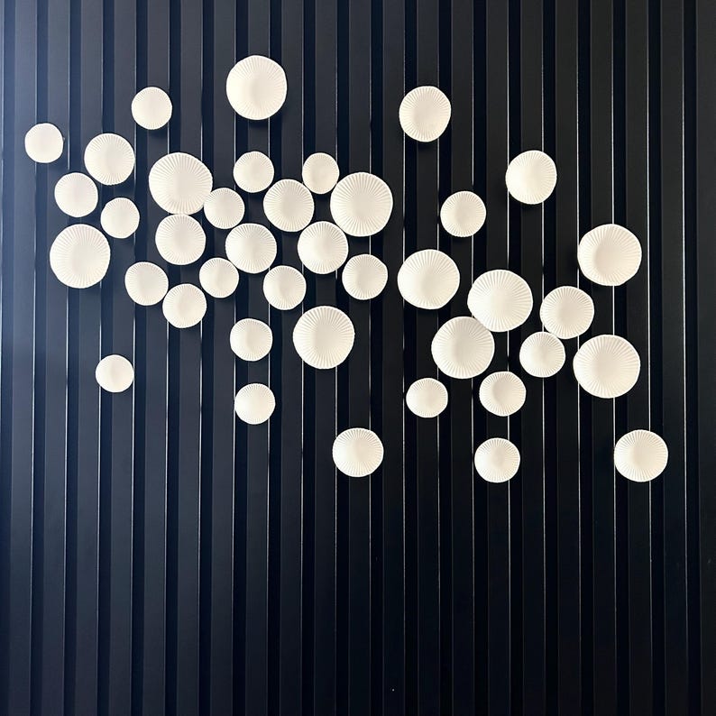 Puede incluir: Una instalaci&oacute;n de arte mural que presenta numerosos elementos decorativos circulares blancos y texturizados de varios tama&ntilde;os. Las piezas est&aacute;n dispuestas sobre un fondo oscuro con l&iacute;neas verticales, creando una est&eacute;tica moderna y minimalista. Las piezas blancas tienen un patr&oacute;n radial.