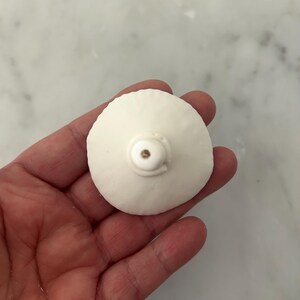 Puede incluir: Un objeto blanco y redondo con una peque&ntilde;a protuberancia descentrada y un acento met&aacute;lico. El objeto se sostiene en una mano, con un fondo claro. Parece ser un elemento decorativo o un componente peque&ntilde;o.