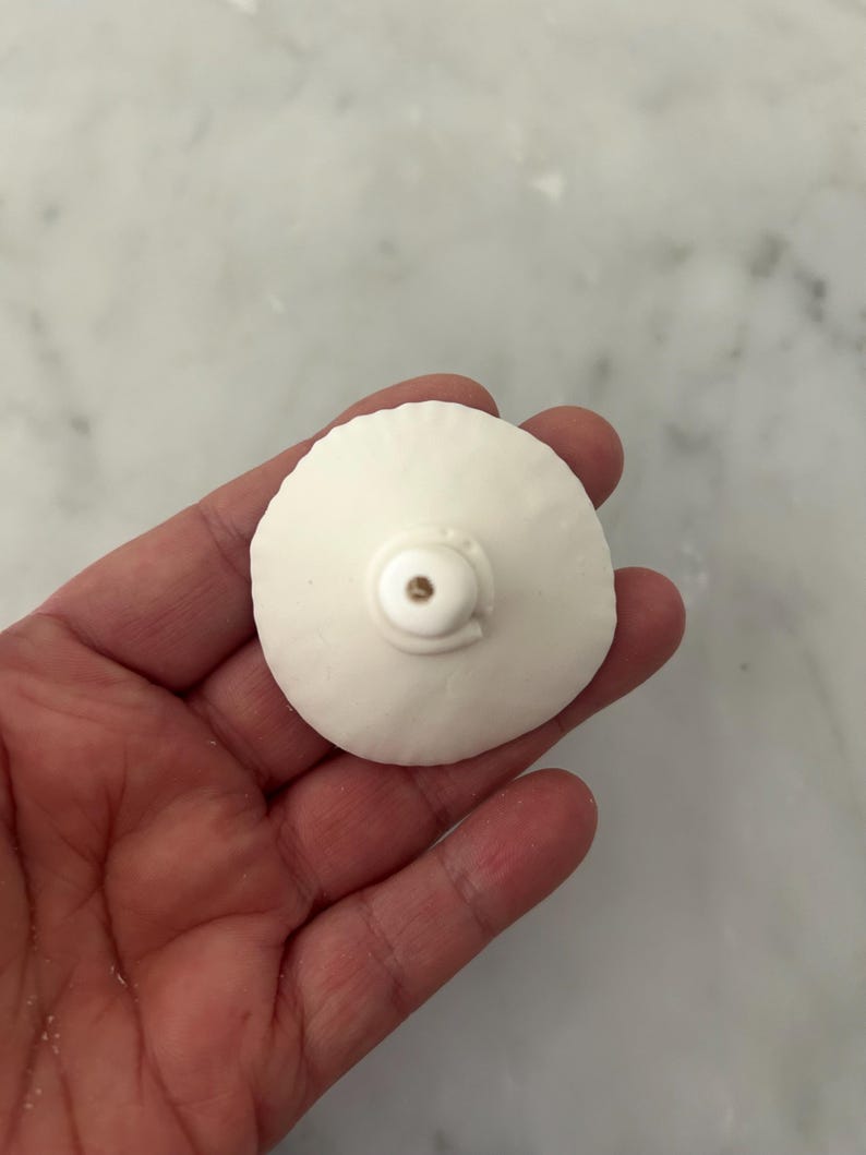 Puede incluir: Un objeto blanco y redondo con una peque&ntilde;a protuberancia descentrada y un acento met&aacute;lico. El objeto se sostiene en una mano, con un fondo claro. Parece ser un elemento decorativo o un componente peque&ntilde;o.