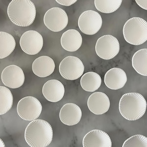 Puede incluir: Vista cenital de numerosos platos peque&ntilde;os, blancos y redondos con textura. Cada plato presenta un patr&oacute;n radial que emana del centro. Est&aacute;n dispuestos sobre una superficie gris claro veteada, creando una composici&oacute;n dispersa y atractiva.