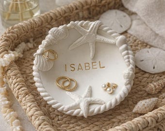 May include: A white, seashell-themed trinket dish with the name "ISABEL" in gold lettering. The dish is decorated with starfish and seashells, and holds gold rings and pearl earrings. It sits in a woven tray with other beach-themed decor.