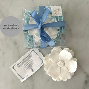 May include: White flower wedding favors in a clear box tied with a light blue ribbon. The box contains shredded paper and a white flower. A single flower and a certificate of authenticity are also visible. The text "personalized wedding favors" is in a gray circle.