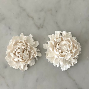 May include: Two white ceramic peonies, each with a delicate, layered design. The peonies are arranged side-by-side on a white marble surface.