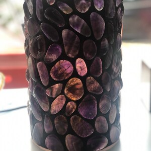 May include: A cylindrical amethyst stone vase with a dark background. The vase is covered in irregularly shaped amethyst stones, showcasing shades of purple and translucent areas. The base has a copper-coloured trim.