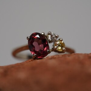 May include: A gold ring with a large, oval-cut, deep red gemstone as the main feature. It's flanked by smaller white and yellow accent stones. The ring is set against a neutral backdrop.