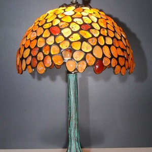 May include: A table lamp featuring a shade made of orange and amber-toned stones. The lamp has a bronze-coloured base and stem with a textured, tree-like design. The lamp is plugged into an electrical outlet.