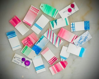 May include: A collection of 15 watercolor gift tags with various designs, including stripes, polka dots, and a Christmas tree. The tags are all white with different colored watercolor designs.