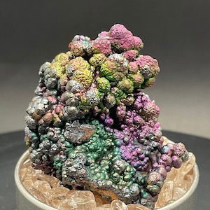 May include: A cluster of iridescent, metallic, green, purple, and silver goethite crystals in a small, silver container. The crystals are arranged in a random, organic pattern.