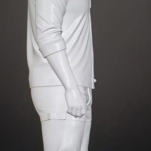 May include: A monochromatic white figurine of a person standing in profile. The figure wears a collared button-down shirt with rolled sleeves, trousers, and boots. The sculpture features a smooth, glossy finish and is presented against a dark background.