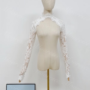 May include: A white lace bolero with long sleeves and a high mock neck collar. The garment features an intricate floral pattern throughout the lace fabric, with scalloped edges along the neckline and cuffs, displayed on a beige mannequin torso.