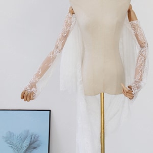 May include: A white lace bridal veil and matching lace sleeves are displayed on a mannequin. The sleeves are long and fitted, with intricate floral lace patterns. The veil is long and flowing, with a delicate lace trim.