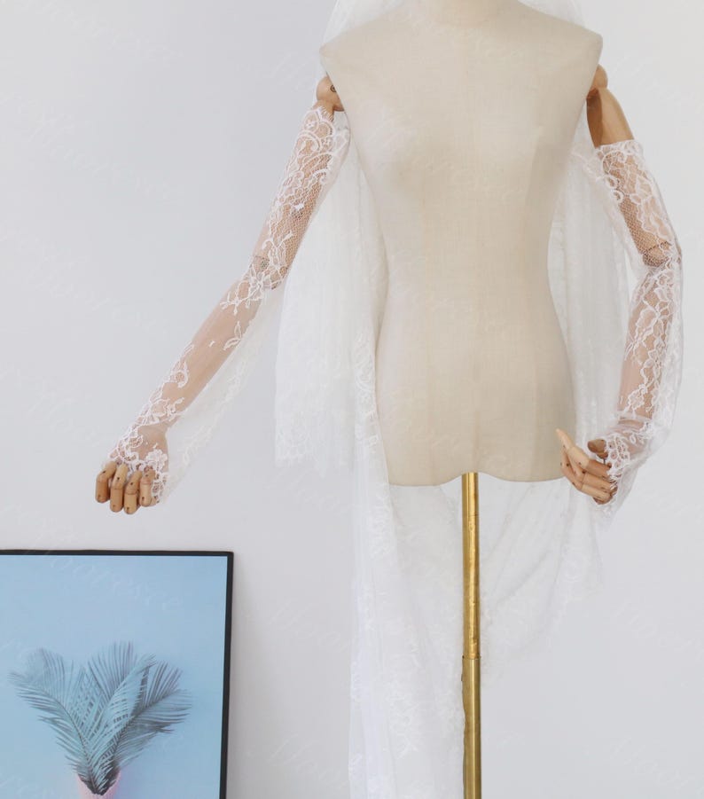 May include: A white lace bridal veil and matching lace sleeves are displayed on a mannequin. The sleeves are long and fitted, with intricate floral lace patterns. The veil is long and flowing, with a delicate lace trim.