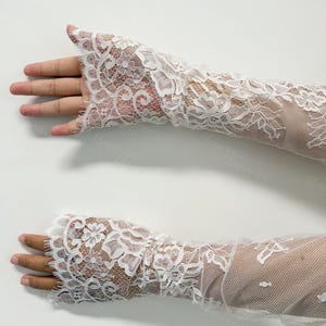 May include: A pair of white lace arm warmers. The arm warmers are crafted from delicate, floral-patterned lace. The lace is detailed and covers the entire arm, extending from the wrist to the mid-forearm. The background is a plain white surface.