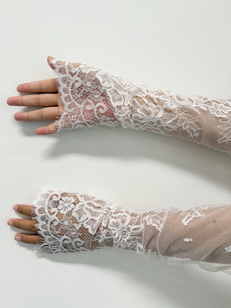 May include: A pair of white lace arm warmers. The arm warmers are crafted from delicate, floral-patterned lace. The lace is detailed and covers the entire arm, extending from the wrist to the mid-forearm. The background is a plain white surface.