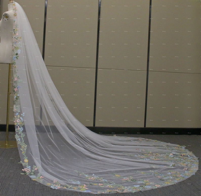 May include: A long, white tulle wedding veil with a floral embroidered border. The embroidery features colorful flowers in shades of pink, blue, and green.
