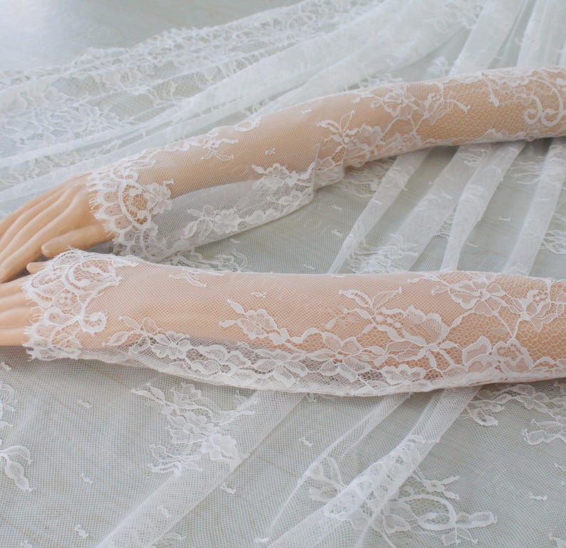 May include: Elegant white lace gloves with floral designs. The gloves are long, reaching up the arm, and have detailed lace edging. The fabric is sheer, creating a soft, romantic look. The gloves are a beautiful accessory.